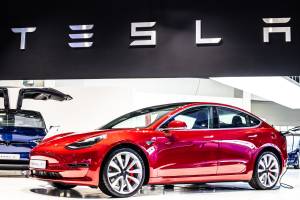 Apresentação do Model 3 em uma feira de carros. O veículo é vermelho e há um banner da Tesla na parte superior da imagem