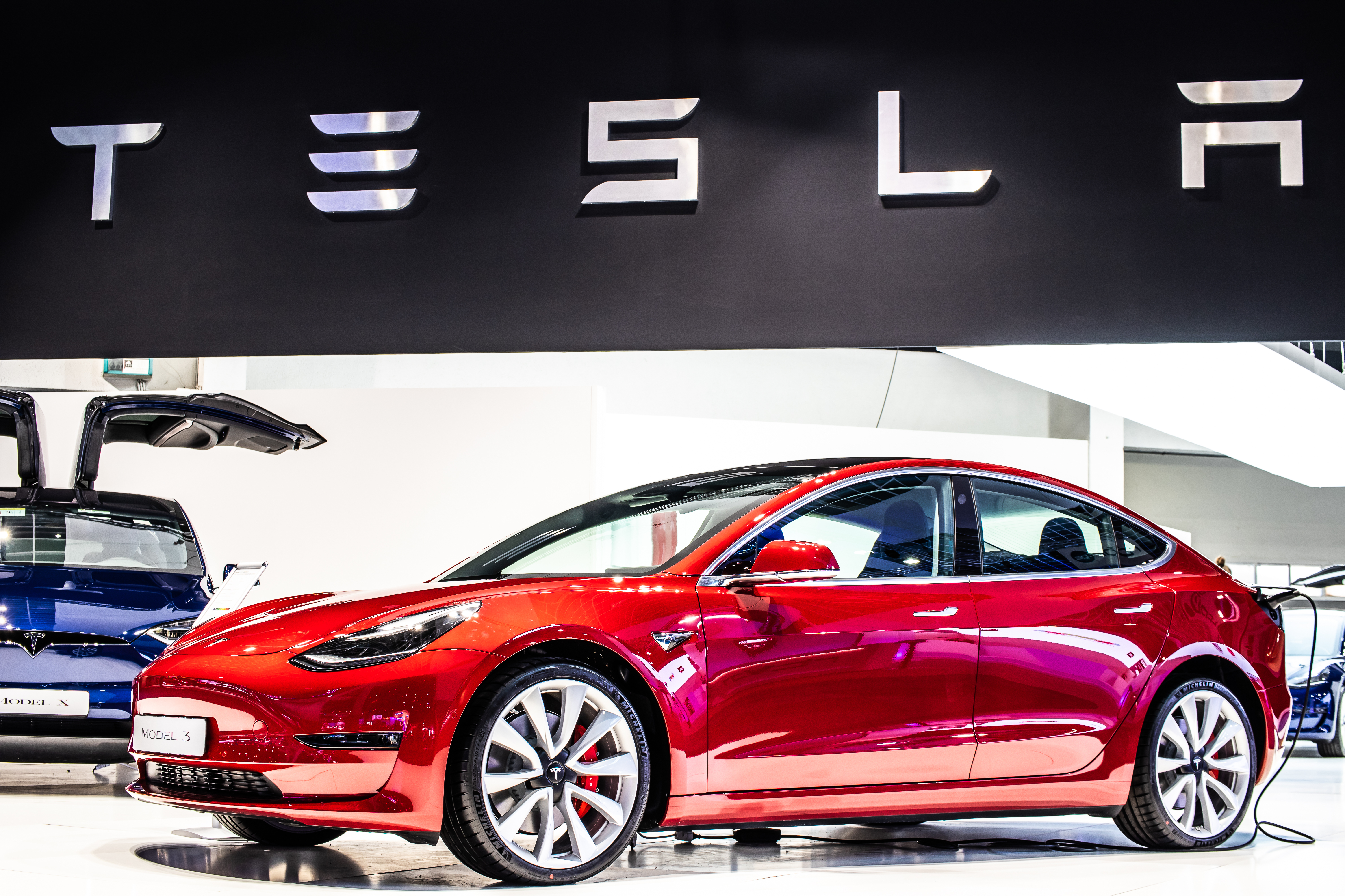 Apresentação do Model 3 em uma feira de carros. O veículo é vermelho e há um banner da Tesla na parte superior da imagem