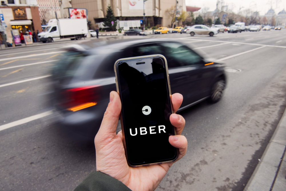 Mão segurando um celular com o logo da Uber centralizado na foto. Um carro passa no fundo da imagem