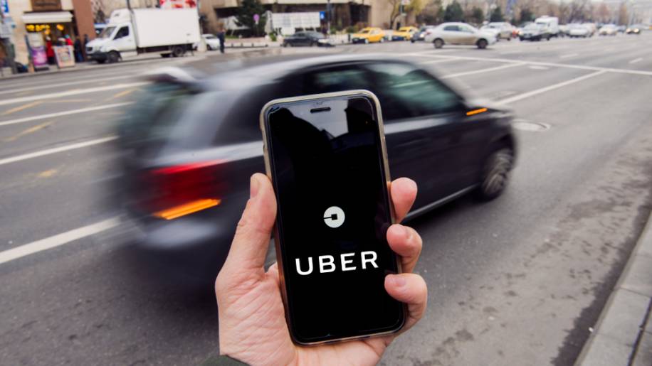 Mão segurando um celular com o logo da Uber centralizado na foto. Um carro passa no fundo da imagem