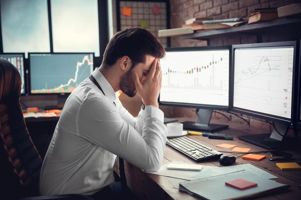 Homem segura o rosto com as mãos, cabisbaixo, diante do computador com gráficos de ações