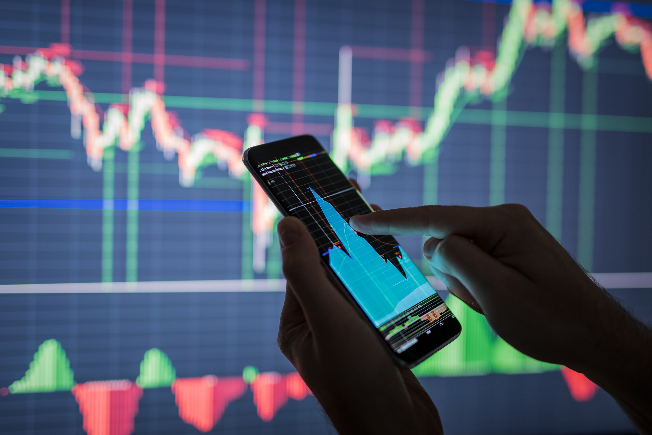 Mão segura um celular e consulta um gráfico em frente a um painel de movimentação de ações em Bolsa - mercado fracionário