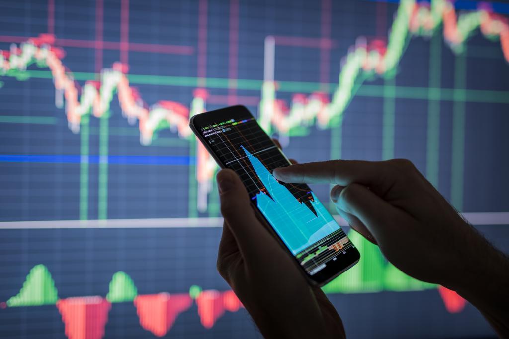 Mão segura um celular e consulta um gráfico em frente a um painel de movimentação de ações em Bolsa
