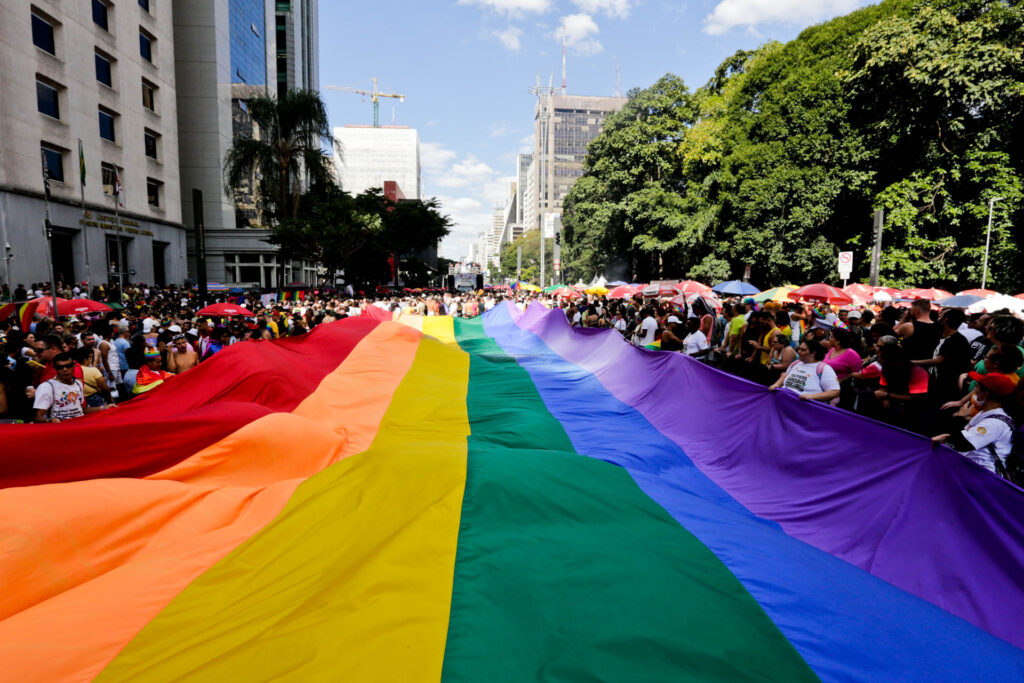 Bandeira com as cores do arco-íris esticada no meio da multidão durante a Parada do Orgulho LGBT+ de SP em 2023