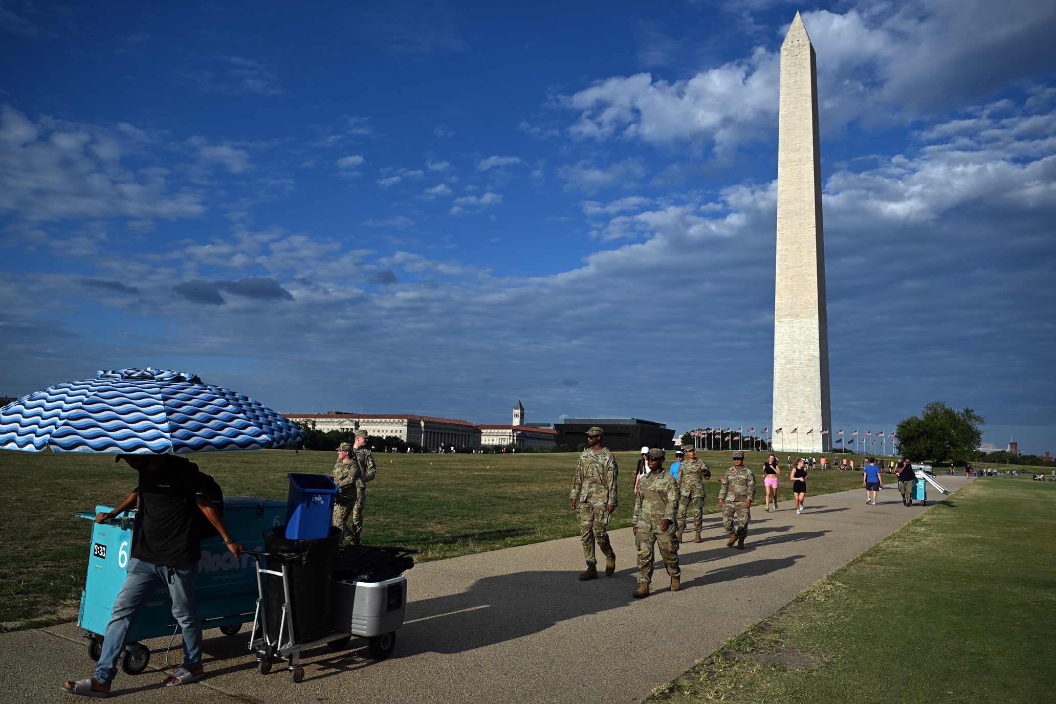 Um vendedor de sorvete é seguido por tropas da Guarda Nacional em patrulha no Monumento a Washington, em Washington, na noite de terça-feira, 12 de agosto de 2025 (Kenny Holston/The New York Times)